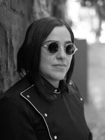 Josette Melchor in round clear-frame sunglasses, leaning on a wall.