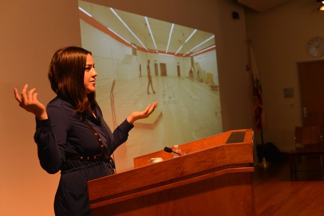 Josette Melchor speaking at a podium, gesturing, with an architectural rendering projected behind her.