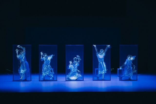 Five dancers inside glass cases rendered as glowing blue wireframe figures against a dark stage.